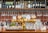 A bar setting featuring a polished gold beer tap with various labeled handles. Behind the bar, there are shelves stocked with bottles of wine and spirits, along with neatly hanging wine glasses. The countertop displays several clean glasses and there are elements of green plants and fruit adding color to the scene.