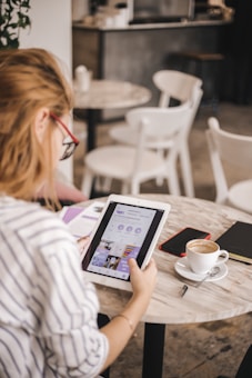 A person with long hair and glasses is seated at a round table in a cafe, using a tablet device that displays a social media profile. The table also holds a cup of coffee, a smartphone, a notebook, and a pen. The background features several white chairs and tables, and a plant is partially visible.