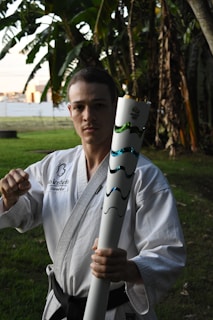 A person wearing a martial arts uniform holds a decorated torch with a firm grip, standing in a grassy outdoor setting surrounded by trees. The individual has a determined expression and is making a fist with their other hand.