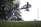 Two martial artists are engaged in a kata in an outdoor setting. One is executing a high flying kick while the other is in a defensive stance on a grass-covered area. Dense greenery and trees are in the background, with the sky visible above.