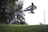 Two martial artists are engaged in a kata in an outdoor setting. One is executing a high flying kick while the other is in a defensive stance on a grass-covered area. Dense greenery and trees are in the background, with the sky visible above.
