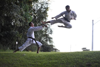 Two martial artists are engaged in a kata in an outdoor setting. One is executing a high flying kick while the other is in a defensive stance on a grass-covered area. Dense greenery and trees are in the background, with the sky visible above.