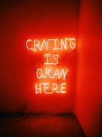 Bright neon text illuminates a dimly lit space, casting a warm orange glow on the surrounding walls. The message conveys a sense of comfort and acceptance.