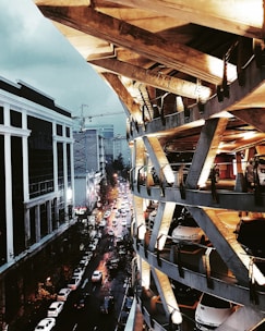 A multi-level parking structure with vehicles is situated next to a busy urban street. The architecture features a unique geometric design with exposed supports and lighting that casts a warm glow on the structure. Adjacent to this, a bustling city street is visible, with several cars and buildings illuminated by evening streetlights, creating a contrast against the dark, cloudy sky.