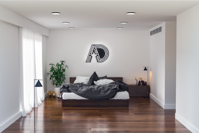A modern bedroom with a minimalistic design features a large bed with dark gray bedding against a white wall. Above the bed, there is a neon-like decorative piece on the wall. On the right, a wooden nightstand holds a small plant and some decor items. A lush green plant is situated to the left near a tall slender lamp, next to a sliding glass door with sheer white curtains. The room has hardwood floors and recessed ceiling lights.