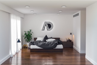 A modern bedroom with a minimalistic design features a large bed with dark gray bedding against a white wall. Above the bed, there is a neon-like decorative piece on the wall. On the right, a wooden nightstand holds a small plant and some decor items. A lush green plant is situated to the left near a tall slender lamp, next to a sliding glass door with sheer white curtains. The room has hardwood floors and recessed ceiling lights.