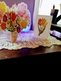 A decorative arrangement sits on a wooden surface covered with a lace doily. There is a three-dimensional card featuring colorful flowers, including roses and tulips, and a separate card displaying an illustration of tulips. A blue diffuser or lamp is visible in the background, softly illuminating the scene with a violet hue.