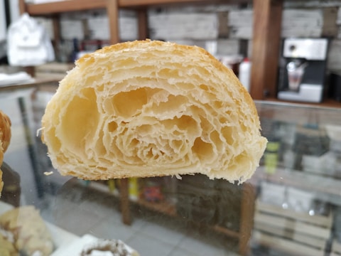 A close-up view of a cross-section of a flaky, golden-brown pastry with visible layers and a soft, airy texture. The pastry is placed on a reflective glass surface in a setting that includes a blurred background with shelves and items.