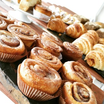 A close-up view of a variety of pastries, prominently featuring cinnamon rolls with a glazed finish. The pastries are arranged on a black slate, with a wooden surface visible. In the background, more baked goods are displayed, including croissants and small cakes.