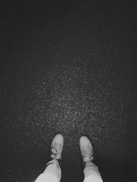 Two feet are visible in light-colored sneakers standing on a dark, textured surface, possibly asphalt.