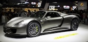 A sleek, silver sports car is displayed on a rotating platform at an auto show. The car attracts a large crowd of onlookers, and its shiny, aerodynamic body reflects the ambient lighting. The stage and backdrop feature branding and digital displays.