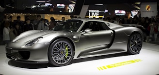 A sleek, silver sports car is displayed on a rotating platform at an auto show. The car attracts a large crowd of onlookers, and its shiny, aerodynamic body reflects the ambient lighting. The stage and backdrop feature branding and digital displays.
