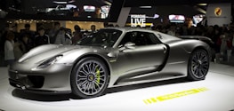 A sleek, silver sports car is displayed on a rotating platform at an auto show. The car attracts a large crowd of onlookers, and its shiny, aerodynamic body reflects the ambient lighting. The stage and backdrop feature branding and digital displays.