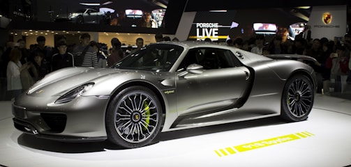 A sleek, silver sports car is displayed on a rotating platform at an auto show. The car attracts a large crowd of onlookers, and its shiny, aerodynamic body reflects the ambient lighting. The stage and backdrop feature branding and digital displays.