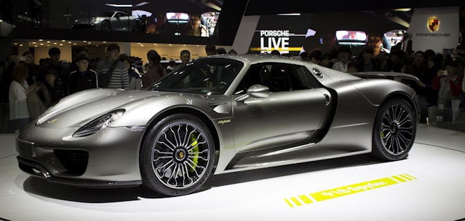 A sleek, silver sports car is displayed on a rotating platform at an auto show. The car attracts a large crowd of onlookers, and its shiny, aerodynamic body reflects the ambient lighting. The stage and backdrop feature branding and digital displays.