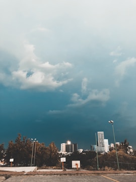 A cityscape is visible in the background, with several tall buildings under a dramatic, cloudy sky. The foreground shows a basketball court surrounded by trees and tall light poles, with a concrete ground.