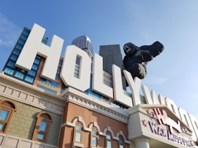 A large building facade with bold white letters spelling 'HOLLYWOOD' prominently displayed. A giant gorilla sculpture is climbing over the letters. The architectural style includes various window designs and textures with a backdrop of skyscrapers.