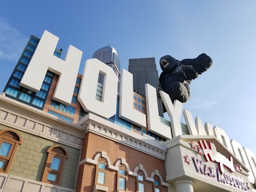 A large building facade with bold white letters spelling 'HOLLYWOOD' prominently displayed. A giant gorilla sculpture is climbing over the letters. The architectural style includes various window designs and textures with a backdrop of skyscrapers.