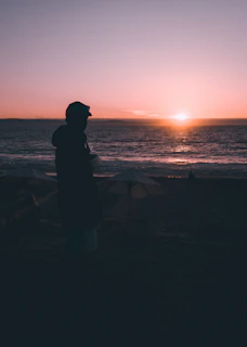 A sunset silhouette of a person holding a large floral umbrella at the beach.