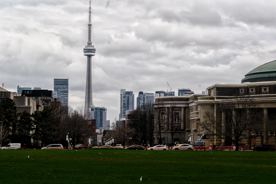 The image depicts a cityscape with a tall, gray-colored tower dominating the skyline. The tower is situated on the left side of the frame and stands out against the cloudy sky. In the foreground, there are several buildings, including one with a green roof that adds a pop of color to the scene.

On the right side of the image, there's a large park filled with lush green grass. The park is dotted with trees, providing a natural contrast to the urban environment. Cars can be seen parked along the street in front of the buildings, indicating a bustling city life.