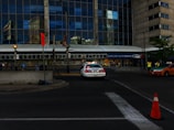 A police car is parked near a building with a large glass facade reflecting nearby structures. People are walking along the sidewalk, and there is a traffic cone and a taxi on the street. A sign above the entrance reads 'Season Ticket Member Entrance.'