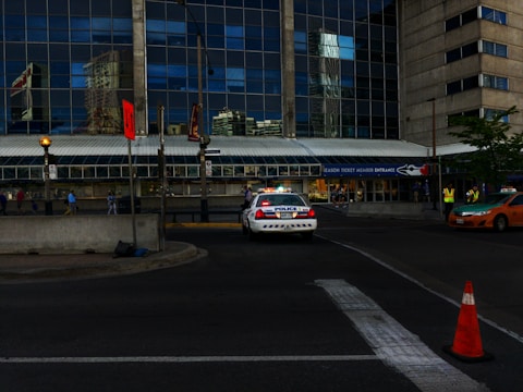 A police car is parked near a building with a large glass facade reflecting nearby structures. People are walking along the sidewalk, and there is a traffic cone and a taxi on the street. A sign above the entrance reads 'Season Ticket Member Entrance.'