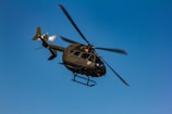 A helicopter is flying in the sky, displaying a military-style design with a dark green body. The rotors are blurred, indicating motion against a clear blue sky.