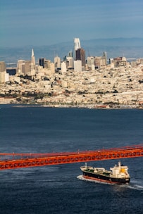 A large cargo ship is traveling beneath a red suspension bridge over a vast body of water. In the background, a sprawling cityscape with numerous skyscrapers and buildings stretches across the horizon, set against a backdrop of distant hills under a clear blue sky.