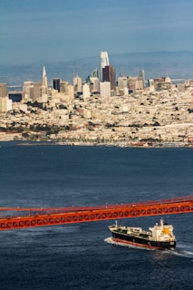 A large cargo ship is traveling beneath a red suspension bridge over a vast body of water. In the background, a sprawling cityscape with numerous skyscrapers and buildings stretches across the horizon, set against a backdrop of distant hills under a clear blue sky.