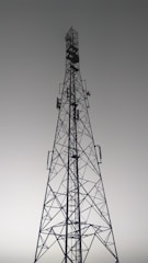 A tall metal communication tower with an intricate lattice structure reaching up towards a gray, overcast sky. Various satellite dishes and antennas are mounted along its frame.