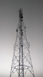 A tall metal communication tower with an intricate lattice structure reaching up towards a gray, overcast sky. Various satellite dishes and antennas are mounted along its frame.