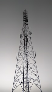 A tall metal communication tower with an intricate lattice structure reaching up towards a gray, overcast sky. Various satellite dishes and antennas are mounted along its frame.
