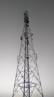 A tall metal communication tower with an intricate lattice structure reaching up towards a gray, overcast sky. Various satellite dishes and antennas are mounted along its frame.
