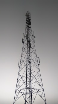 A tall metal communication tower with an intricate lattice structure reaching up towards a gray, overcast sky. Various satellite dishes and antennas are mounted along its frame.