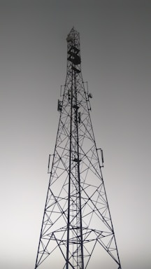 A tall metal communication tower with an intricate lattice structure reaching up towards a gray, overcast sky. Various satellite dishes and antennas are mounted along its frame.