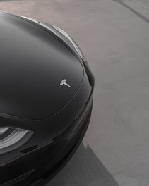 A close-up view of the front corner of a sleek electric car displaying a smooth, dark hood with a logo. The car is parked on a light gray, flat surface, possibly a parking lot or driveway. The design is modern and elegant.