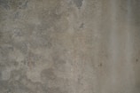 The image consists of a rough, textured concrete surface with an uneven pattern and subtle color variations. Scattered patches and streaks are visible, possibly from wear or environmental exposure.