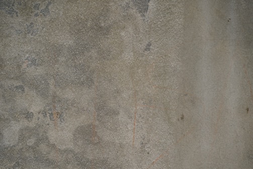 The image consists of a rough, textured concrete surface with an uneven pattern and subtle color variations. Scattered patches and streaks are visible, possibly from wear or environmental exposure.
