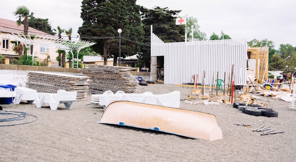 A construction site with scattered materials including wooden planks, plastic chairs, and tires. A white building with a red cross sign is partially constructed in the background, surrounded by debris and tools. Trees and a residential building are visible in the background.