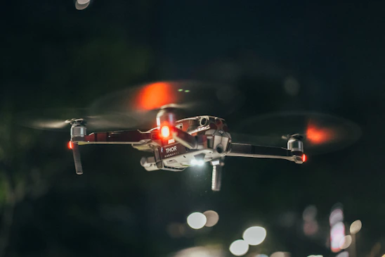 A sleek FPV drone flying through a neon-lit cityscape at dusk, with blue and orange lights reflecting off glass buildings.