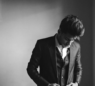 A person dressed in a formal suit, looking down with a contemplative expression. The image is in black and white, focusing on the details of the suit's texture and the person's hairstyle.