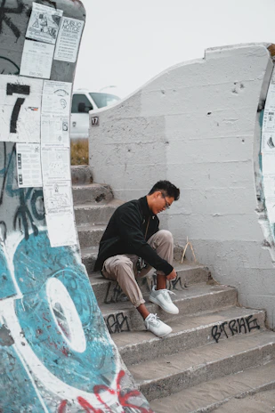 A young person seated on concrete steps wearing an original, unbranded jacket and slim-fit joggers against a graffiti wall.