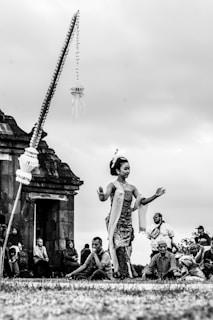 A traditional dancer performs outdoors with an ancient stone structure in the background. The dancer, wearing cultural attire, gracefully moves amidst a gathering of people who are seated and watching the performance. The sky appears overcast, creating a serene ambiance.