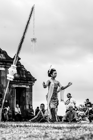 A traditional dancer performs outdoors with an ancient stone structure in the background. The dancer, wearing cultural attire, gracefully moves amidst a gathering of people who are seated and watching the performance. The sky appears overcast, creating a serene ambiance.