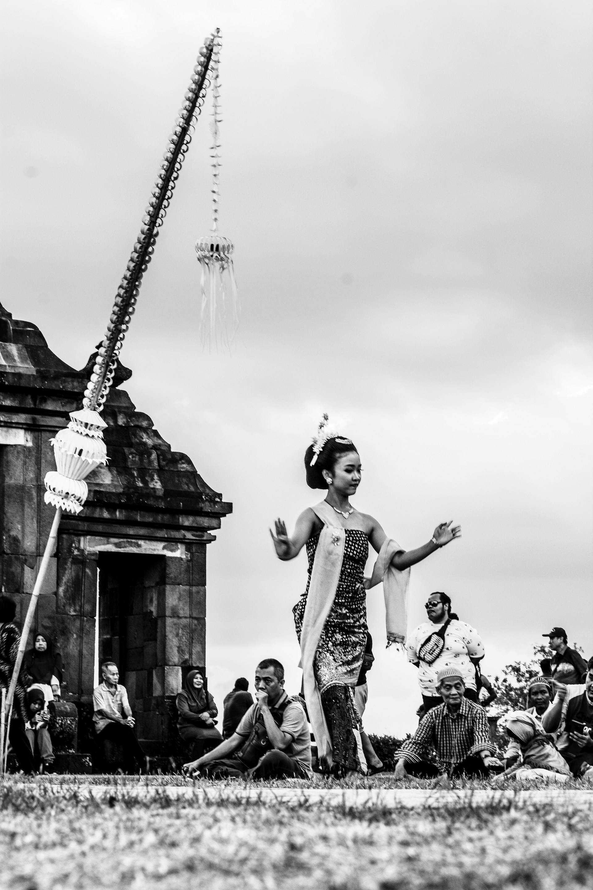 A traditional dancer performs outdoors with an ancient stone structure in the background. The dancer, wearing cultural attire, gracefully moves amidst a gathering of people who are seated and watching the performance. The sky appears overcast, creating a serene ambiance.