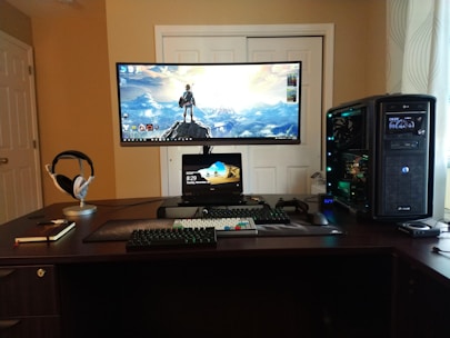A well-organized computer setup on a dark wooden desk featuring a large curved monitor displaying a scenic desktop wallpaper, a smaller tablet or laptop positioned beneath, a mechanical keyboard, gaming mouse, and large tower PC with visible internal components. Headphones rest on a stand, and a closed notebook with a pen lies next to it.