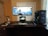 A well-organized computer setup on a dark wooden desk featuring a large curved monitor displaying a scenic desktop wallpaper, a smaller tablet or laptop positioned beneath, a mechanical keyboard, gaming mouse, and large tower PC with visible internal components. Headphones rest on a stand, and a closed notebook with a pen lies next to it.
