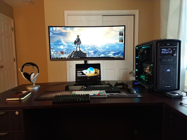 A well-organized computer setup on a dark wooden desk featuring a large curved monitor displaying a scenic desktop wallpaper, a smaller tablet or laptop positioned beneath, a mechanical keyboard, gaming mouse, and large tower PC with visible internal components. Headphones rest on a stand, and a closed notebook with a pen lies next to it.