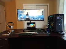 A well-organized computer setup on a dark wooden desk featuring a large curved monitor displaying a scenic desktop wallpaper, a smaller tablet or laptop positioned beneath, a mechanical keyboard, gaming mouse, and large tower PC with visible internal components. Headphones rest on a stand, and a closed notebook with a pen lies next to it.