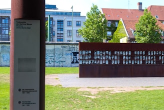 A memorial installation with a metal structure displaying numerous black-and-white portraits, set against the backdrop of a building with a red-tiled roof and a section of the graffiti-covered Berlin Wall. A plaque with inscriptions is also visible in the foreground, providing information about the victims of the Berlin Wall.